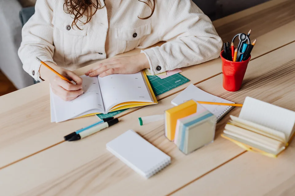 Hands writing with a pencil in an open notebook at a wooden desk, with books, loose papers and a red cup of pens nearby.