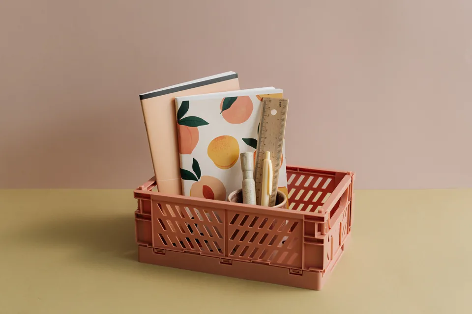 A coral basket with school notebooks, pens and a ruler on a warm neutral tabletop.