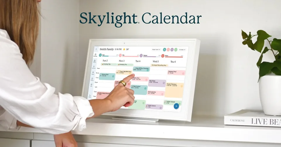A hand tapping a white-framed Skylight touchscreen on a shelf, showing a color-coded week of family events.
