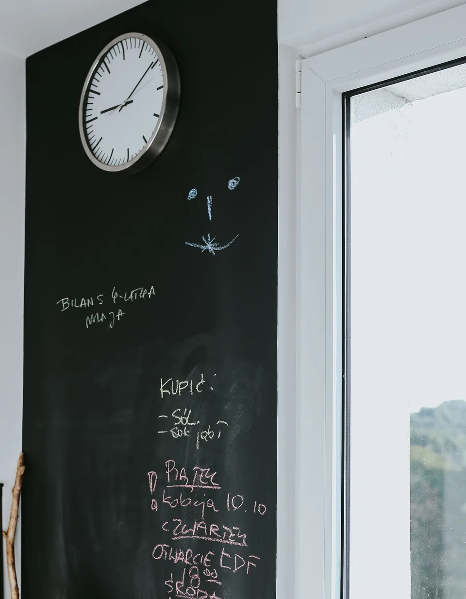 A kitchen chalkboard wall with a round clock, a chalk smiley face and a handwritten shopping list and weekday plan beside a window.