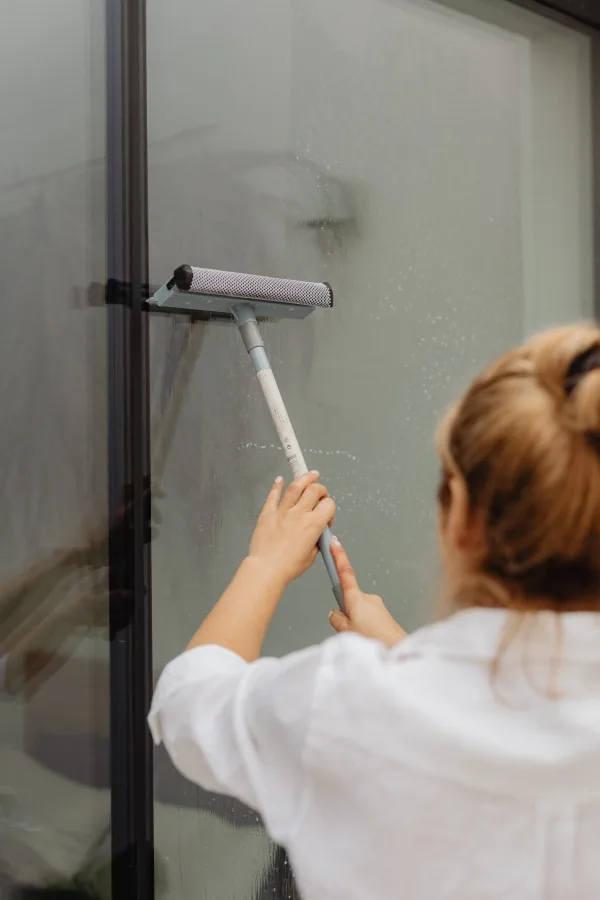 Back view of a person wiping a tall glass window with a long-handled cleaning tool.