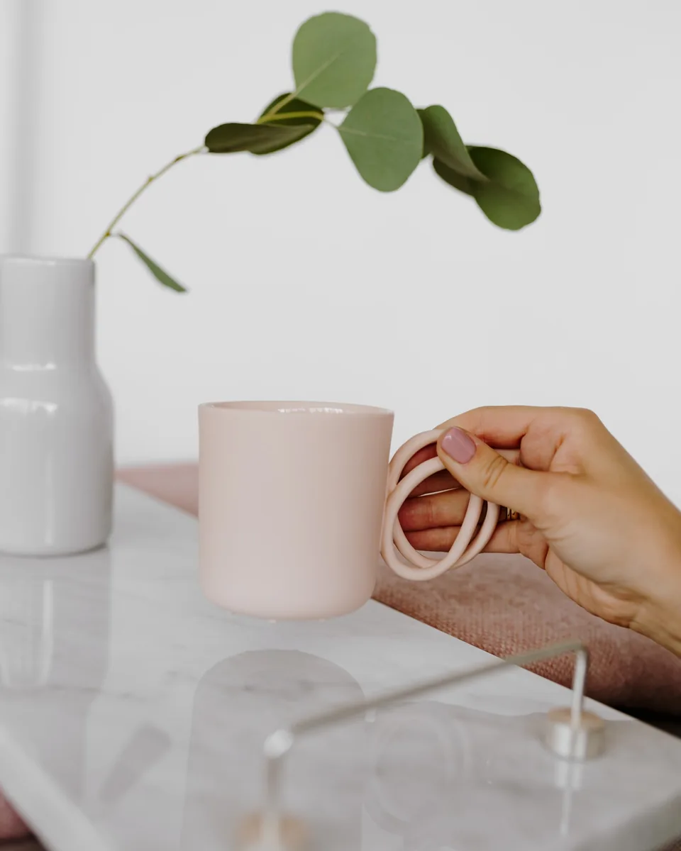 A hand holding a pale pink mug beside a white vase with a sprig of eucalyptus, on a marble tray.