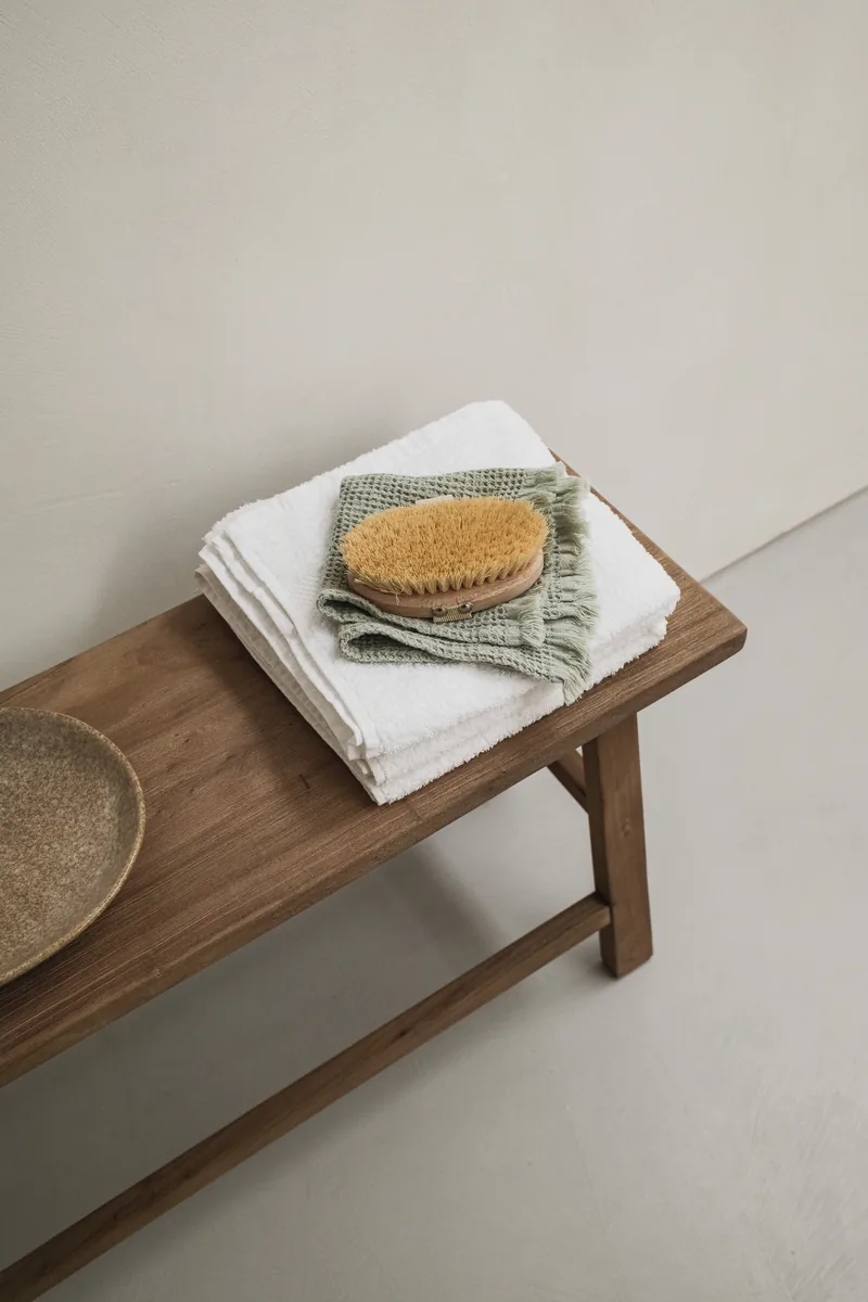 Folded white and sage towels with a wooden body brush on a simple wooden bench.