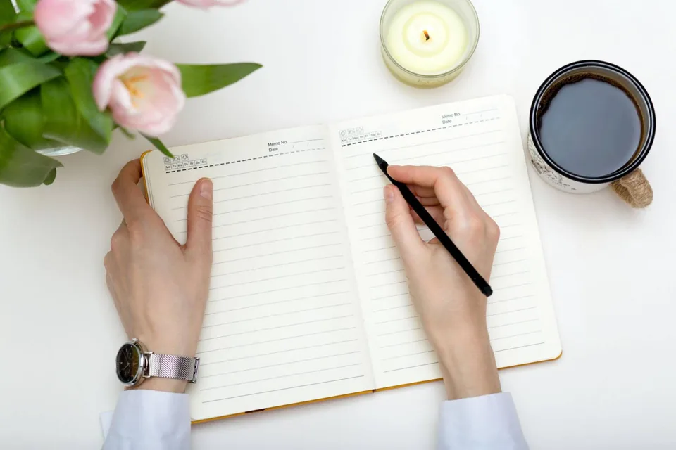 Two hands hold an open lined notebook and a pencil on a white table, beside a mug of black coffee, a lit candle and pink tulips.